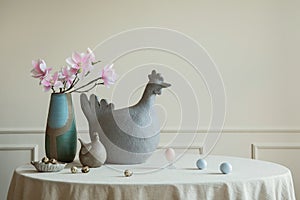 Minimalist composition of easter dining room interior with copy space, gray hen sculpture, colorful eggs, vase with magnolia,
