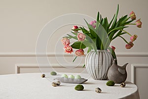 Minimalist composition of easter dining room interior with round table, vase with tulips, bowl with colorful eggs, hen sculpture,