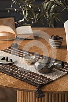 Minimalist composition of living room interior with wooden round table, book, incense, bowl with nuts, patterned table pad, beige