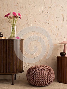 Minimalist composition of living room interior with wooden sideboard, glass vase with pink flowers, lamp, casket, pouf, books,