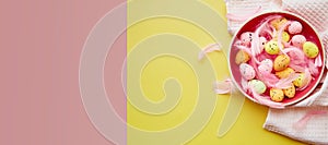 Minimalist Easter colorful eggs in bowl and pink feathers on yellow background. Extra wide banner. Copy space