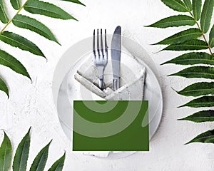 Minimalist mock wedding or  birthday table setting with silver cutlery, marble plate, vintage napkin and tropical leaves on white