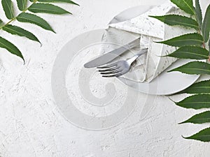 Minimalist mock wedding or  birthday table setting with silver cutlery, marble plate, vintage napkin and tropical leaves on white