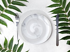 Minimalist table setting with marble plate, cutlery, tropical leaves on white background.