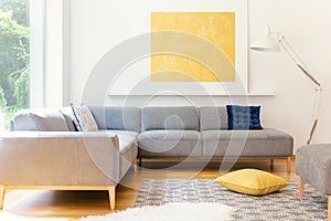 A minimalist, yellow poster and a white, industrial floor lamp in a sunny living room interior with a patterned rug and vibrant de