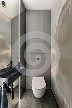 Minimalistic composition of bathroom interior with white toilet seat, gray tiles, big mirror, dark towel, simple sink, light on