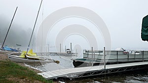 A Minnesota lake beach on a foggy morning with boat docks, a pontoon, and beached sailboats waiting to be enjoyed. Tourism resort