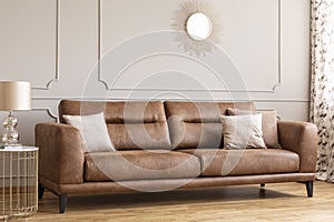 Mirror above leather couch with pillows in grey living room interior with gold lamp on table