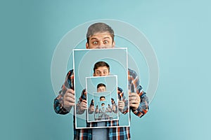 Mirror effect. Young man in casual clothes holding his portrait isolated on light blue background. Human emotions.