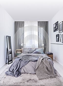 Mirror next to bed with grey sheets in white bedroom interior with drapes at window. Real photo