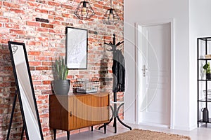 Mirror next to wooden cabinet in entrance hall interior with white door and poster on red brick wall