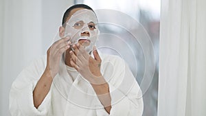 Mirror POV of young African American man in white bathrobe putting on purifying facial mask standing in bedroom in the