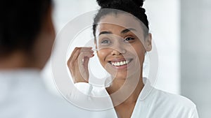 Mirror reflection African American woman cleaning ears, using cotton bud