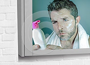 Mirror reflection portrait of young confused attractive man using beauty facial beauty product applying cream and reading lotion b