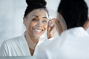 Mirror reflection smiling African American woman plucking eyebrows with tweezers