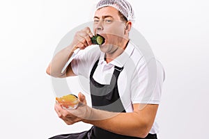 A mischievous chef in a black robe tastes a cucumber on a white plate with a knife in his hands and a bowl of vegetables