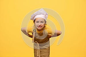 Mischievous little girl in chef's hat and apron, grimacing, making faces, posing with hands on waist, isolated on