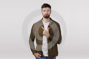 Miserable and lonely young sad man with beard, wearing coat, pulling gloomy unhappy expression as pointing upper left