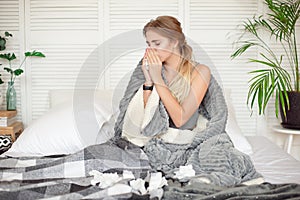 Miserable young woman sitting on the bed wrapped in warm blanket feeling sick with flu