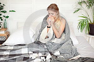 Miserable young woman sitting on the bed wrapped in warm blanket feeling sick with flu