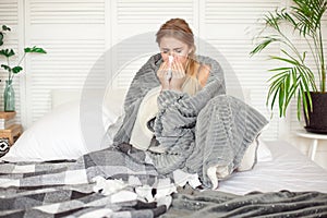 Miserable young woman sitting on the bed wrapped in warm blanket feeling sick with flu