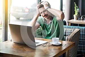 Mistake. Young sad businessman in green t-shirt sitting and working on laptop and holding his head because he made mistake