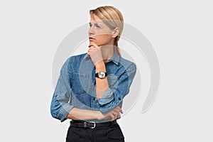 Mistrustful pensive young business woman in denim shirt with finger touching her chin and looking to the one side over white