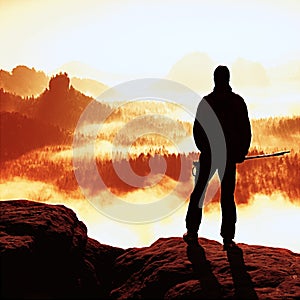 Misty day in rocky mountains. Silhouette of tourist with poles in hand. Hiker stand on rocky view point above misty valley.