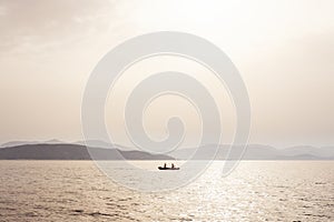 Misty mood seascape with fishermen boat in the middle of the sea and coastline in background under fog sunlight