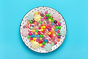 mix colorful sweets - white round plate with black peas, lollipop, meringue, chocolate, sprinkle on blue background Flat lay Top