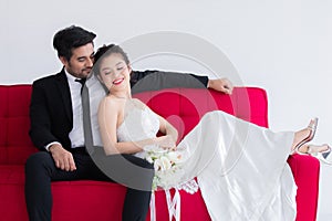 Mix race wedding concept, Beautiful Asian woman lie down on chest of Caucasian handsome man on red sofa bed, happy couple spent
