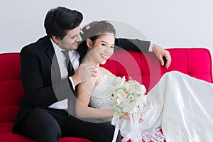 Mix race wedding concept, Beautiful Asian woman lie down on chest of Caucasian handsome man on red sofa bed, happy couple spent