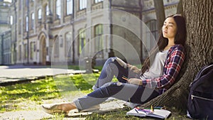 Mixed female relaxing under tree with eyes shut, stressful day, peacefulness