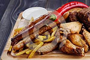 Mixed fresh grilled meat and sausage with pepper, french fries and sauce on wooden cutting board. Assorted meat with vegetables