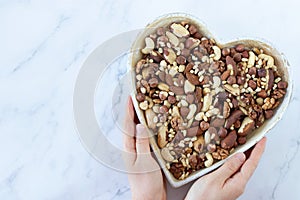 Mixed nuts (hazelnut, almond, walnut, Brazil, and pine nuts) in a heart-shaped dish with female hands isolated on white