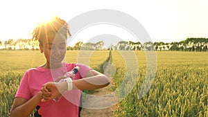 Mixed race African American girl teenager female young woman runner using smart watch and running on path through field at sunset