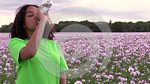 Mixed race African American girl teenager female young woman walking in field of pink poppy flowers drinking a bottle of water