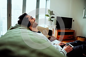 Mixed race business man working from home smiling and laughing listening to music with headphones sitting on comfy couch