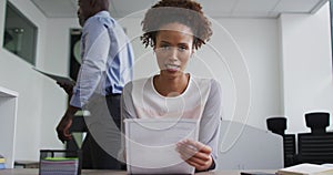 Mixed race businesswoman sitting at desk, holding documents and talking during video call in office