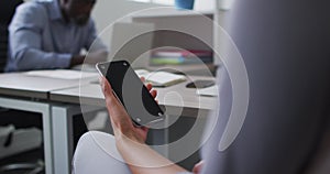 Mixed race businesswoman sitting at desk and using smartphone with copy space on screen in office