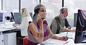 Mixed race businesswoman sitting using computer talking on phone headset in office