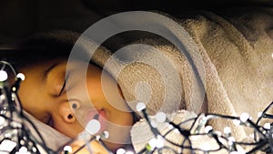 Mixed-race child is sleeping in Christmas lights. Blurred and retouched.