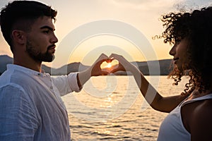 Mixed race couple, Caucasian man with her Hispanic girlfriend, forming a heart with their hands framing the setting sun on a lake