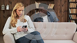 Mixed race couple sitting on a couch while husband is trying to peep into wife's mobile phone as she is typing a text