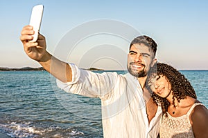 Mixed race couple in vacation at sea resort making a self portrait at sunset or dawn - Handsome smiling bearded man take photo