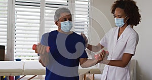 Mixed race female physiotherapist wearing mask helping senior exercise using dumbbells