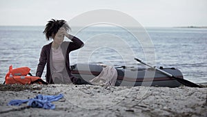 Mixed race girl sitting on beach near boat, flood disaster victim, shipwreck