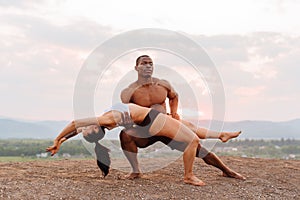 Mixed race gymnastic couple with perfect bodies in sportswear dancing on mountains landscape background. Pink sunset sky