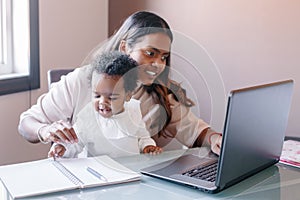 Mixed race Indian mother with African black baby working online from home on Internet. Workplace of freelancer woman with kid.