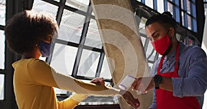 Mixed race male cafe worker wearing face mask taking temperature of female coworker